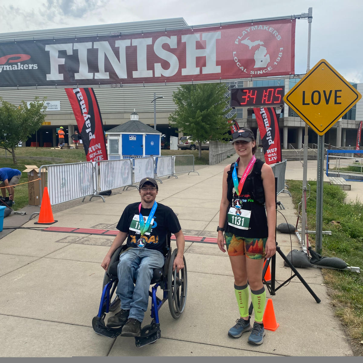 Two runners at the finish line of a race, one standing and smiling at the camera, and the other sitting in a wheelchair, smiling at the camera.